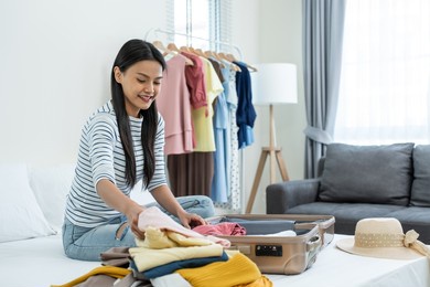 asian young beautiful woman preparing clothes and packing a suitcase. attractive female tourist traveler feel happy and relax while preparing luggage on bed, ready to travel on holiday vacation trip.