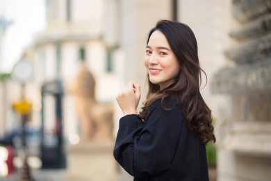 portrait of a beautiful long-haired asian female in a black coat, carrying a walking bag, smiling in a good mood in the city outdoors.