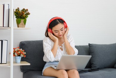 young asian woman wear red headphones relaxing and listening the music while using laptop computer on couch in living room, girl enjoys audio sound, relaxing