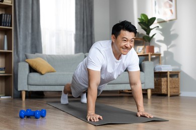 cheerful middle aged asian man in homewear exercising alone at home, planking on fitness mat, looking at copy space and smiling, enjoying morning workout, cozy living room interior