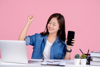 happy asian businesswoman hold mobile phone with blank empty screen doing winner gesture isolated on a pink background