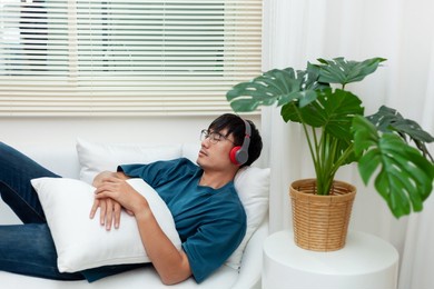 the idea of ​​relaxing at home. a handsome asian man wears headphones while lying on the sofa listening to music in his apartment. meditation or podcast with eyes closed. relax in the bedroom