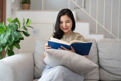 happy asian young woman sitting on sofa and reading book at home -people and leisure concept