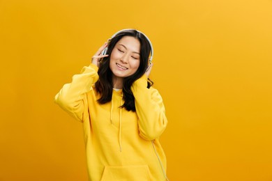 enjoyed favorite songs asian student young woman in yellow hoodie sweatshirt sings closing eyes posing isolated on over orange studio background. cool offer. perfect playlist sound app concept