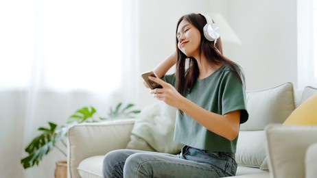 young asian woman listening to music on couch in living room at home. happy asia female using mobile smartphone, wearing headset and sitting on sofa