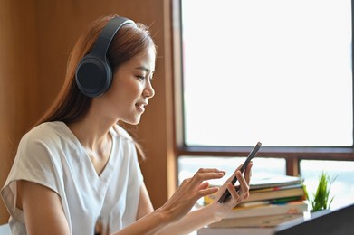beautiful millennial asian female university student listening to music with her modern wireless headphones and using smartphone.