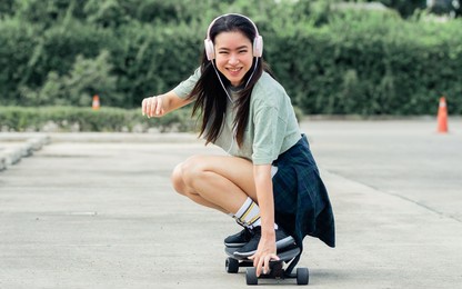beautiful sportive hipster happy asian woman wearing casual shirt and shorts with headphone to listen music while playing skateboard as hobby in free time day on holiday, smiling at outdoor park