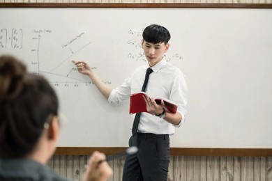 an asian male teacher in a classroom near the whiteboard is writing and teaching a lesson with a special lecture in a class column. math teacher explains arithmetic sums to university students.