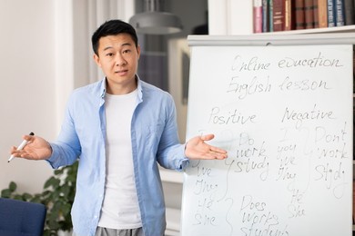 chinese male teacher standing near whiteboard teaching english standing in modern classroom. tutor explaining language grammar rules talking looking at camera. education concept
