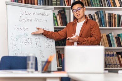 asian male teacher teaching online having remote class via video call on laptop computer standing near whiteboard at workplace. internet technology and education. selective focus