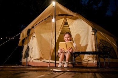 cheerful and happy millennial asian female camping at the campground, sits in front of her camping tent alone and enjoys reading a novel at night.