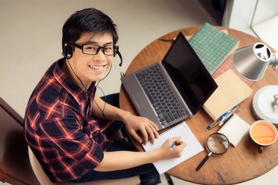 young asian man in a headset working on laptop and making notes, view from above