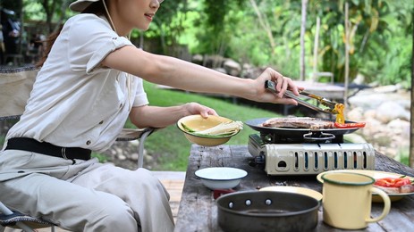 female traveler siting on folding chair and grilling barbecue during camping near the river bank. outdoor activity or vacation concept