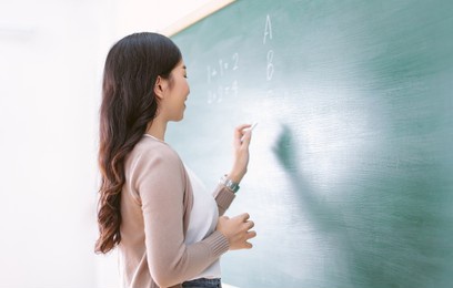 side view of smiling female asian teacher writing on chalkboard, teach english to students in classroom. happy woman writing on blackboard wall during math class. back to school concept