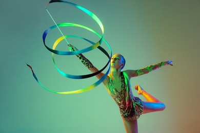 young girl, female rhythmic gymnast training with multicolored ribbon isolated over green background in neon light. concept of action, motion, sport, motivation, competition. copyspace for ad