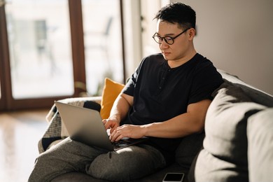 adult calm asian man sitting on sofa with laptop and working at home