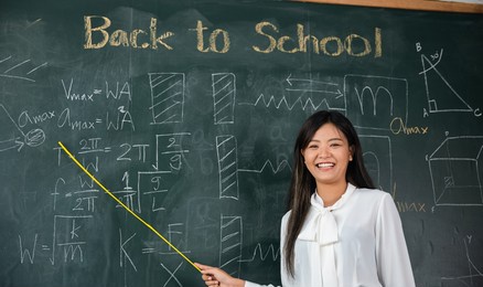 back to school concept. asian female teacher smiling with wooden stick pointing to blackboard at school in classroom, happy beautiful young woman standing hold pointer to back board