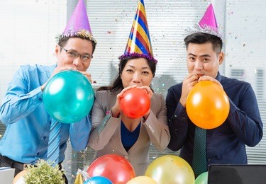 three asian business colleagues blowing up balloons and looking at the camera