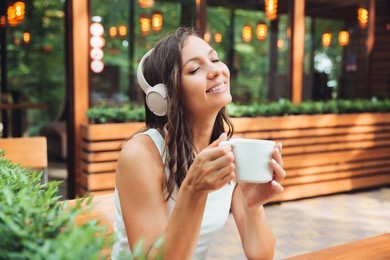 a young beautiful woman with headphones is sitting at a table in a summer cafe and drinking coffee or tea. generation z.