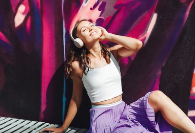 a young happy  woman dressed in a white top and purple skirt, headphones, listening to music, dancing on a city street near the wall of a building. the concept of the lifestyle of urban youth people.