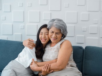 portrait of happy asian senior, mother or grandparent white hair embracing her beautiful daughter or grandchild smiling and closing eyes with feel love while sit on grey couch in living room at home.