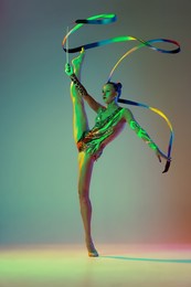 flexible young girl, female rhythmic gymnast training with ribbon isolated over green background in neon light. concept of action, motion, sport life, motivation, competition. copyspace for ad