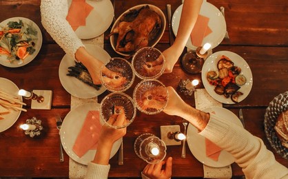 happy friends clinking champagne glasses while celebrating new years eve or christmas, sitting at festive dinner table with traditional food and burning candles. top view