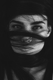 portrait of a mysterious woman with her long brown hair wrapped in strands across her face looking at the camera with seductive luminous eyes, monochrome head and shoulders image