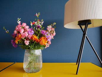 colorful garden flowers in glass vase on yellow sideboard against petrol blue wall with stylish table lamp. high quality photo