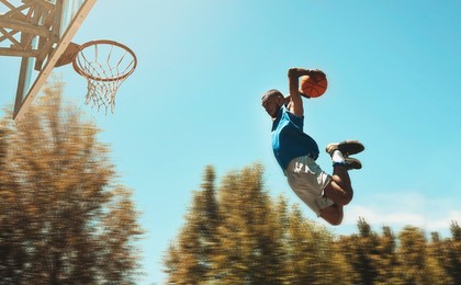 basketball, sports and dunk with a man athlete jumping or flying through the air to score while playing on an outdoor court against the sky. sport, fitness and exercise with a male basketball player