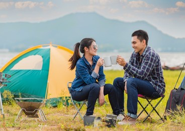 happy asian young couple sitting on picnic chair drinking tea and coffee while tent camp lakeside at parks outdoors on vacation holiday. adventure lifestyle of man and woman with camping in nature.