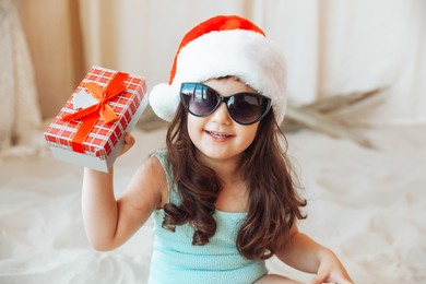 a little girl is sitting on the sand in a santa hat. the child smiles and looks at the gift box. merry christmas. winter holidays. space for copying.