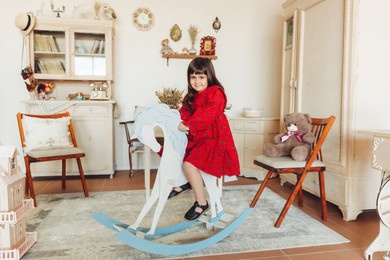 a charming little girl in a dress made of natural material rides a rocking horse in her room. antique nursery interior