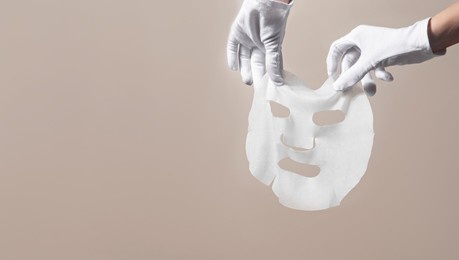 female gloved hands holding white facial mask sheet on beige background. beauty skincare concept