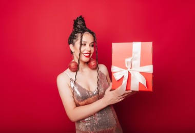 a woman in a red christmas hat with dreadlocks holds a gift on a red background. isolate. a woman in a smart dress. new year christmas
