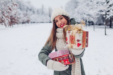 happy woman holding christmas presents gift boxes in snowy winter park offering one. festive holiday season. space. girl wearing warm knitted clothes and red lipstick