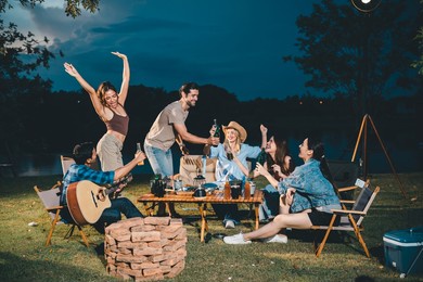young hipster people having fun to music dining and drinking together in campsite - travel vacation lifestyle and youth culture concept, happy friends group toasting beers at barbecue camping party