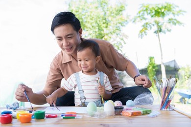father and baby little son painting eggs with brush in class workshop. happy asian dad embracing his little son and teaching painting eggs with water colour