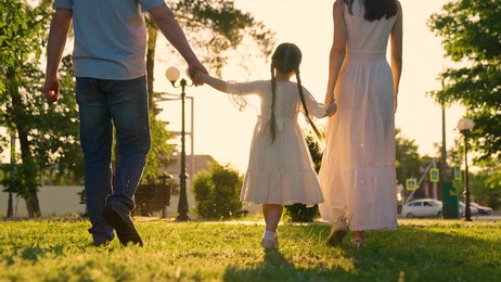 family, child walking hand in hand in city park on green grass in sun. slow motion. active family, mom daughter, dad play together outdoors. happy childhood, future of child. family walk concept, day