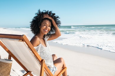 portrait of happy african american woman sunbathing on wooden deck chair at tropical beach while looking at camera. smiling black girl enjoying vacation at seaside with copy space. woman relaxing.