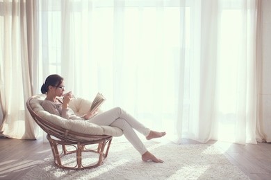 young woman at home sitting on modern chair in front of window relaxing in her living room reading book and drinking coffee or tea