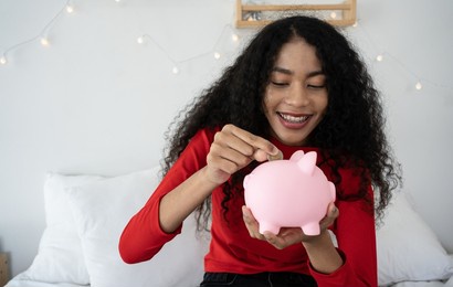 happy black woman puts coin in pink piggy bank. smiling cheerful young woman sitting at home and money box. people and finance, successful business, saving up concept.