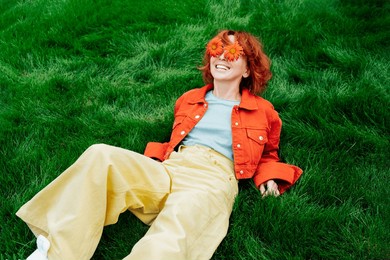 relaxed happy smiling redhead woman with freckles and red orange gerbera flower sunglasses lying on green grass and enjoy the moment. positive vibe emotions. fashion girl. spring, summer mood.