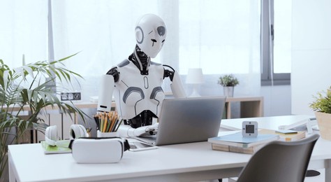 a robot resembling a human sits at a desk in an office, using a laptop to showcase the value of automation in repetitive and menial tasks.