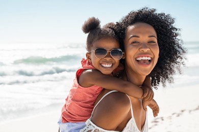 smiling black mother and beautiful daughter wearing sunglasses having fun on the beach. portrait of happy african american woman giving a piggyback ride to her cute little girl wearing shades.