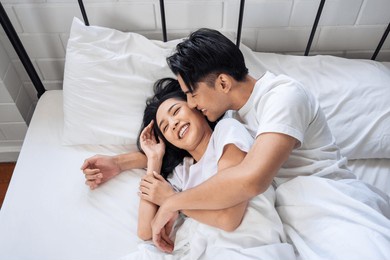 asian husband kissing his wife while lying down sleep on bed in bedroom. beautiful new marriage couple in pajamas feeling happy and relax spending leisure time in the early morning together in house.