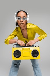 stylish african american woman listening music in wireless headphones while holding boombox isolated on grey