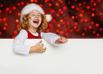 a portrait of a laughing cute little girl in santa hat looking out from behind empty blank with her hands on it, isolated against christmas glitter close up. holidays concept. invitation. funny kids. 