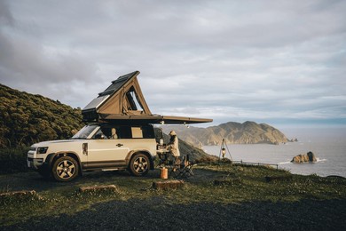 couple sitting in a camping chair in nature with a view of island in a beautiful blue sky with an overlanding car with a rooftop tent and an awning.