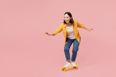 full body young happy fun cool woman of asian ethnicity wear yellow shirt white t-shirt riding skateboard pennyboard isolated on plain pastel light pink background studio portrait. lifestyle concept